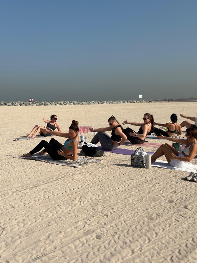 A small Beach Pilates Dubai class mid seated twist on the sand, arms reaching out to the side